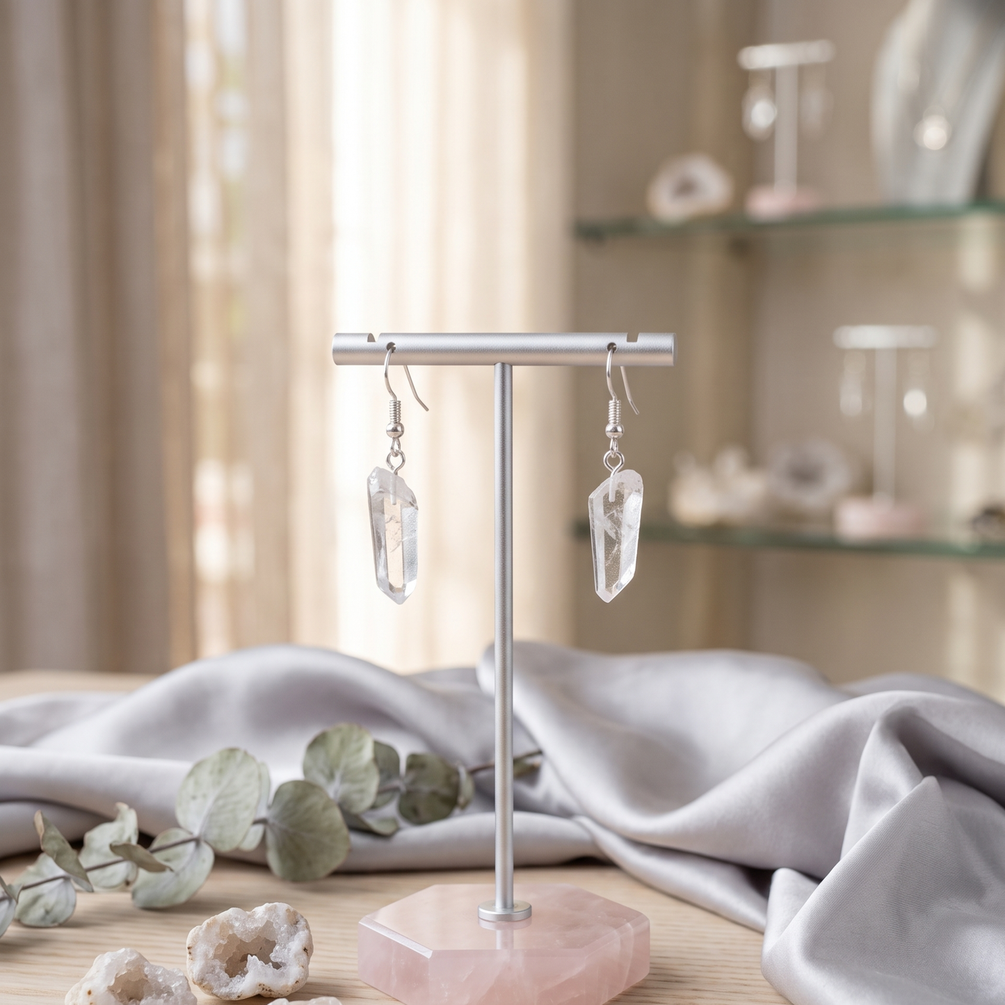 Silver-plated raw clear quartz point earrings hanging on a minimalist T-bar display stand on a elegant wooden dresser with satin fabric accents.